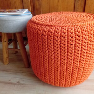 May include: A bright orange crocheted footstool with a round top. The footstool is made of thick yarn and has a textured surface.