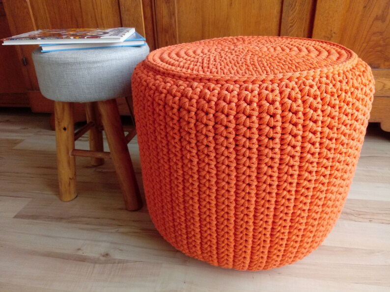 May include: A bright orange crocheted footstool with a round top. The footstool is made of thick yarn and has a textured surface.