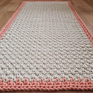 May include: A long, rectangular rug with a white center and a pink border. The rug is made of a woven material and has a textured surface.