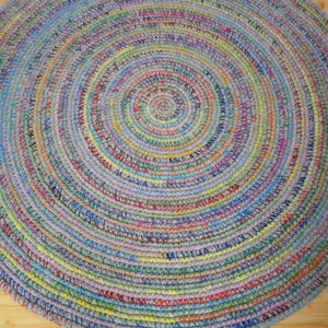 May include: A round, multi-coloured braided rug with a circular pattern. The rug is made of recycled materials and has a variety of colours, including blue, green, yellow, and red.