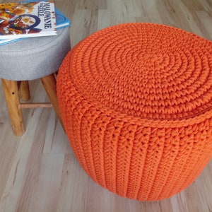 May include: A bright orange crocheted footstool with a circular top and a textured pattern. The footstool is sitting on a wooden floor.