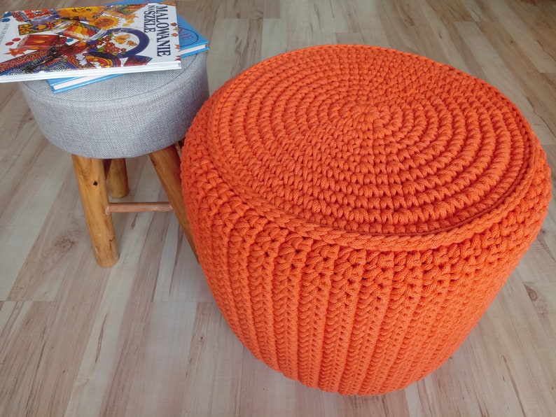 May include: A bright orange crocheted footstool with a circular top and a textured pattern. The footstool is sitting on a wooden floor.