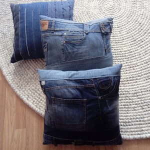 Puede incluir: Tres cojines de mezclilla en varios tonos de azul. Dos cojines están hechos de jeans reciclados, mostrando bolsillos, costuras y cinturillas. El tercer cojín es azul liso con costuras verticales. Los cojines descansan sobre una alfombra redonda de tejido blanco roto.