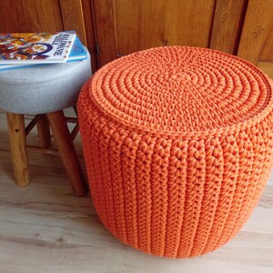 May include: A bright orange crocheted footstool with a circular top and a textured pattern. The footstool is sitting on a wooden floor.