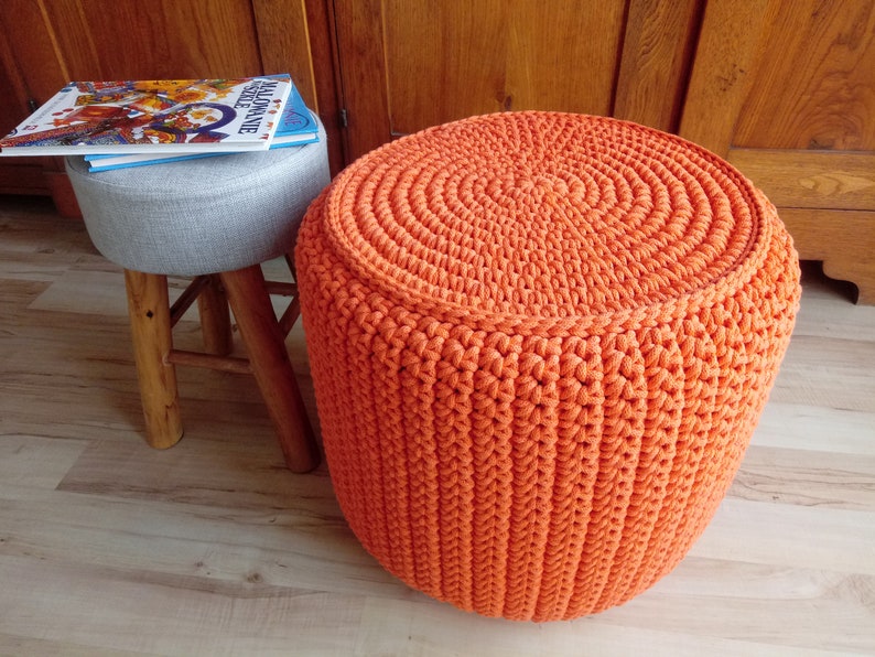 May include: A bright orange crocheted footstool with a circular top and a textured pattern. The footstool is sitting on a wooden floor.