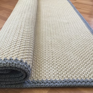 Puede incluir: Una alfombra de crochet de color crema con un borde de crochet gris. La alfombra está enrollada, mostrando la textura del crochet.