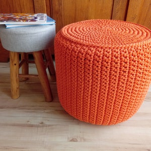 May include: A bright orange crocheted footstool with a round top and a woven texture. The footstool is sitting on a wooden floor.