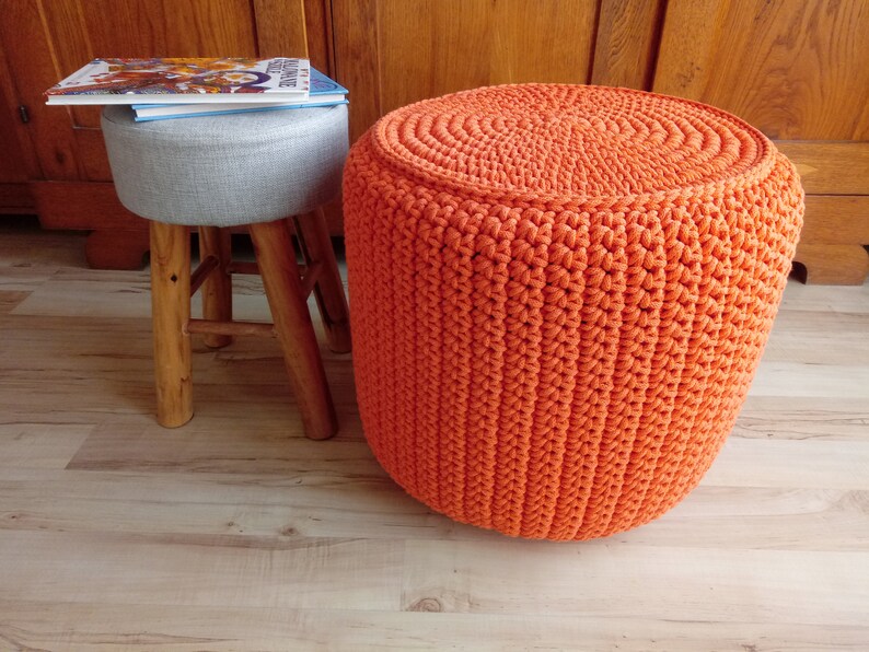 May include: A bright orange crocheted footstool with a round top and a woven texture. The footstool is sitting on a wooden floor.