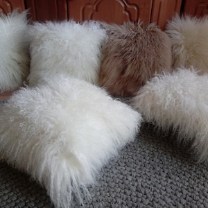 Puede incluir: Cinco almohadas mullidas, cuatro blancas y una marrón, dispuestas sobre una superficie gris texturizada. Las almohadas están hechas de un material suave y peludo.