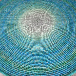 May include: A round, crocheted rug with a gradient pattern of green, blue, and gray. The rug is made of thick yarn and has a textured surface.
