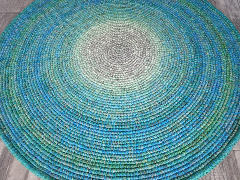 May include: A round, crocheted rug with a gradient pattern of green, blue, and gray. The rug is made of thick yarn and has a textured surface.