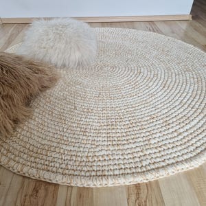 May include: A circular, cream-coloured rug with a concentric circle pattern. Two fluffy, textured cushions in light brown and white are on the rug. The rug is made of woven material and is on a wooden floor.