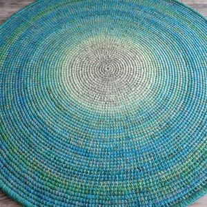 May include: A round, crocheted rug with a gradient of green, blue, and gray colors. The rug is made of a thick, textured yarn and has a circular pattern.