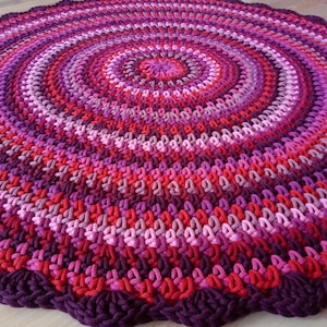 May include: A round, crocheted rug with a multi-colored design. The rug is made of thick yarn in shades of pink, red, and purple.