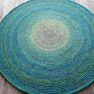 May include: A round, crocheted rug with a gradient of colors from light gray in the center to dark blue on the outer edge. The rug is made of a soft, textured yarn and has a circular shape.