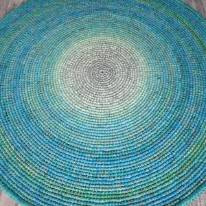 May include: A round, crocheted rug with a gradient pattern in shades of blue, green, and gray. The rug is made of a soft, textured yarn and has a circular shape.