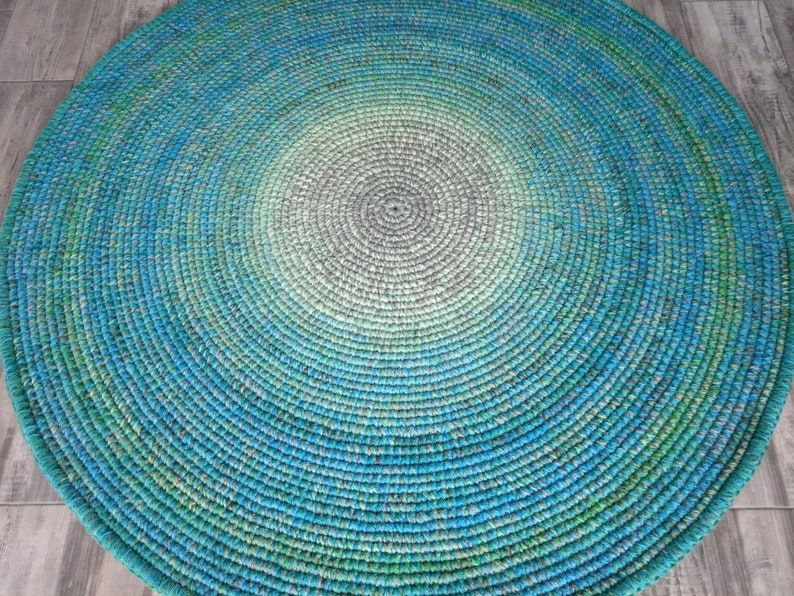 May include: A round, crocheted rug with a gradient pattern in shades of blue, green, and gray. The rug is made of a soft, textured yarn and has a circular shape.