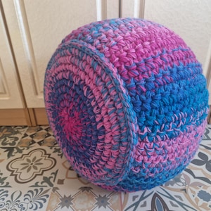 May include: A round, handmade pouf in shades of pink and blue. The crochet design features concentric circles of colour, creating a visually appealing texture. The pouf is a decorative and functional item.