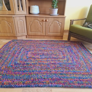 Peut inclure: Un tapis rectangulaire fait à la main avec un design multicolore. Le tapis présente un motif répétitif de bleus, de violets, d'oranges et de verts. Il est posé sur un sol en bois, avec un meuble en bois et un fauteuil vert en arrière-plan.