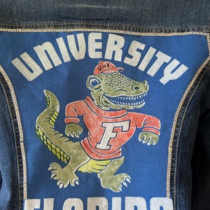 May include: Denim jacket with a blue panel featuring the words "UNIVERSITY FLORIDA" and a cartoon alligator wearing a U of F hat and shirt. The jacket has rhinestone trim.