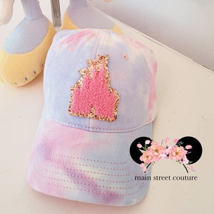 May include: A pastel blue and pink tie-dye baseball cap with a pink glitter castle applique. The text "main street couture" is visible in the bottom right corner of the image.