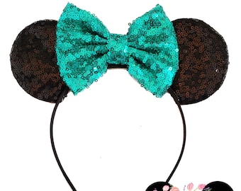 Op de afbeelding: Zwart en turquoise pailletten muis oren haarband met een grote strik. De haarband is versierd met een bloemenontwerp en de tekst "main street couture".