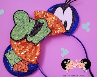 Op de afbeelding: Een paar blauwe en oranje pailletten Minnie Mouse oren met een groen en zwart Goofy-personage op het ene oor en een blauw en wit Goofy-personage op het andere oor. De oren zijn bevestigd aan een blauw hoofdband. Het hoofdband heeft de tekst "main street couture" erop.