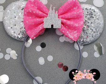 Puede incluir: Orejas de Minnie Mouse de lentejuelas plateadas con un lazo rosa y un dije de castillo de purpurina plateada. La diadema es gris.