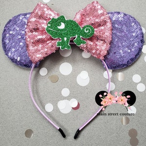 May include: Purple and pink sequined Minnie Mouse ears with a green chameleon charm.  The ears are attached to a purple headband.