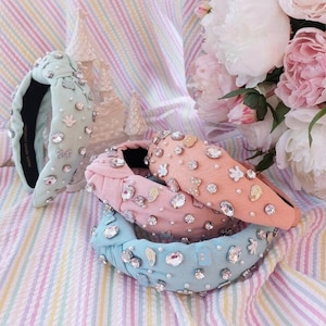 May include: Four colourful headbands with a knotted design, adorned with rhinestones and small decorative elements. The headbands are arranged in a stack on a striped background.