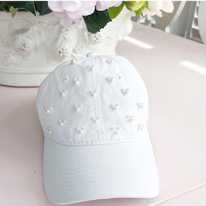 May include: A white baseball cap with a white pearl embellishment design. The cap has a curved brim and a white adjustable strap.