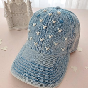 Puede incluir: Gorra de béisbol de mezclilla azul claro decorada con cuentas de perlas blancas con forma de Mickey Mouse. La gorra tiene una visera curva y un botón en la parte superior. Fondo con figura de castillo blanco.