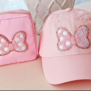 May include: A pink and white polka dot bow is embroidered on a pink baseball cap and a pink zippered pouch.
