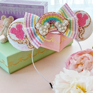 May include: Rainbow and heart-shaped balloon themed Minnie Mouse ears headband with a white headband and a pink, blue, yellow, and green striped bow. The bow has a rainbow with white clouds and pearls.