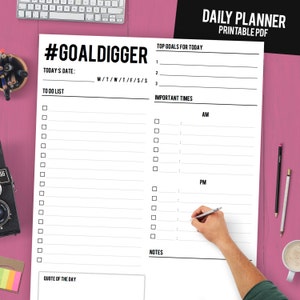 Puede incluir: Un planificador diario blanco con texto negro, incluyendo "#GOALDIGGER" y "DAILY PLANNER PRINTABLE PDF". El planificador tiene secciones para listas de tareas, objetivos principales, horarios importantes y notas. Una mano escribe en el planificador con un bol&iacute;grafo. El fondo es rosa.
