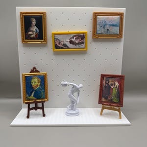 May include: Miniature art display featuring framed paintings and a white sculpture of a discus thrower. The paintings include portraits and landscapes, with gold and brown frames. The display is on a white surface.