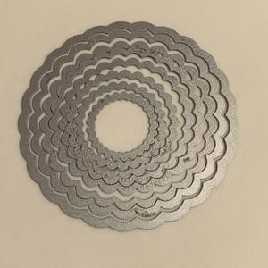 May include: A set of seven nested circular metal cutting dies, each featuring a scalloped edge. The silver-toned dies are arranged concentrically from largest to smallest, creating a layered, ring-like appearance with a textured metallic finish.