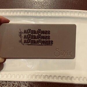 May include: A rectangular, brown die-cutting tool with the words "ALTERATIONS" in English and Spanish, and the Sizzix logo. The tool is held on a white tray.