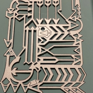 May include: A silver-colored metal die cut featuring an intricate design of arrows, chevrons, and geometric shapes. The die cut has a tribal or Southwestern aesthetic, with various arrowheads and linear patterns. The design is cut out, creating negative space.