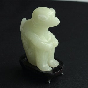 Puede incluir: Una pequeña escultura de jade blanco de un mono sentado sobre una base de madera negra. El mono está tallado en un estilo realista y tiene una expresión serena.