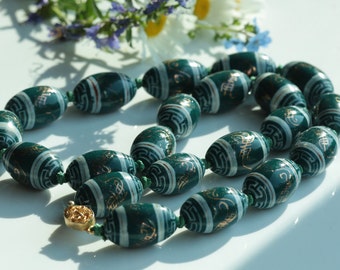 May include: A green and gold beaded necklace with a white background and gold lettering. The beads are oval shaped and have a glossy finish.