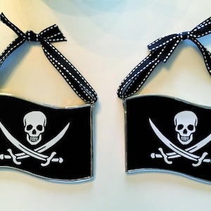 May include: Two black and white pirate flag ornaments with a skull and crossbones design. Each flag is attached to a black ribbon with white polka dots.