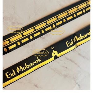 May include: Black ribbon with gold geometric patterns and the words "Eid Mubarak" in gold lettering. The ribbon features a crescent moon and star design. Ideal for gift wrapping or crafting.