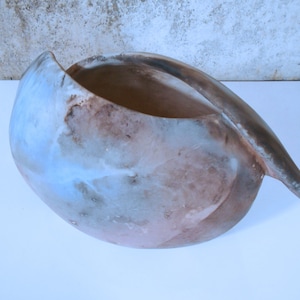 May include: A ceramic bowl with a unique, organic shape. The bowl is glazed in shades of blue, brown, and white, creating a mottled effect. The bowl has a wide opening and a long, thin spout.
