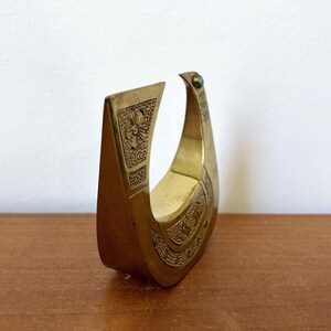May include: A gold-colored metal sculpture of a bird with an ornate design. The bird has a green gemstone on its head.
