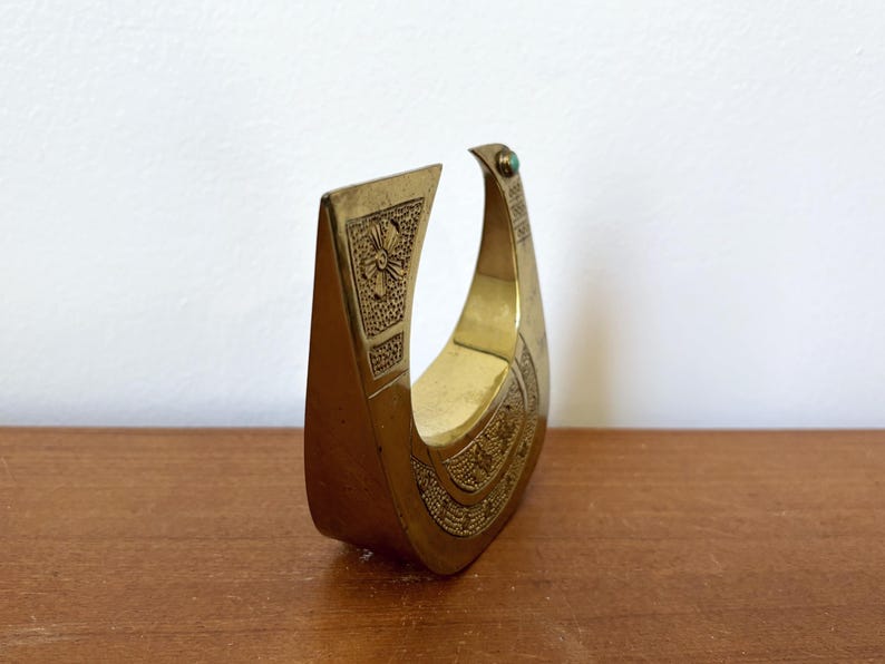 May include: A gold-colored metal sculpture of a bird with an ornate design. The bird has a green gemstone on its head.