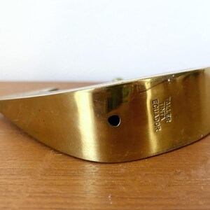 May include: A gold-colored metal object with a curved shape. The object has a hole in the center and the text "TALLER TINTA ECUADOR" is engraved on the side.