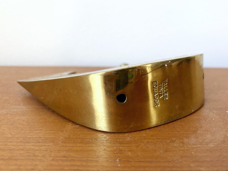 May include: A gold-colored metal object with a curved shape. The object has a hole in the center and the text "TALLER TINTA ECUADOR" is engraved on the side.