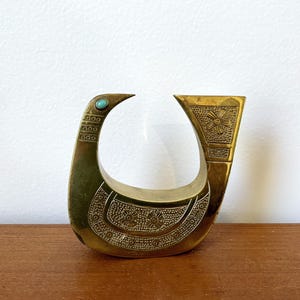 May include: A gold-colored metal bird sculpture with a turquoise stone accent. The bird is stylized and has a geometric design.
