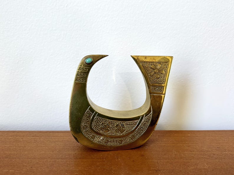 May include: A gold-colored metal bird sculpture with a turquoise stone accent. The bird is stylized and has a geometric design.